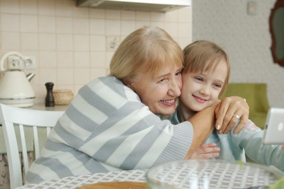 Modern kid girl is taking selfie with loving grandmother using smartphone posing for camera hugging sitting at kitchen table together. Family and gadgets concept.